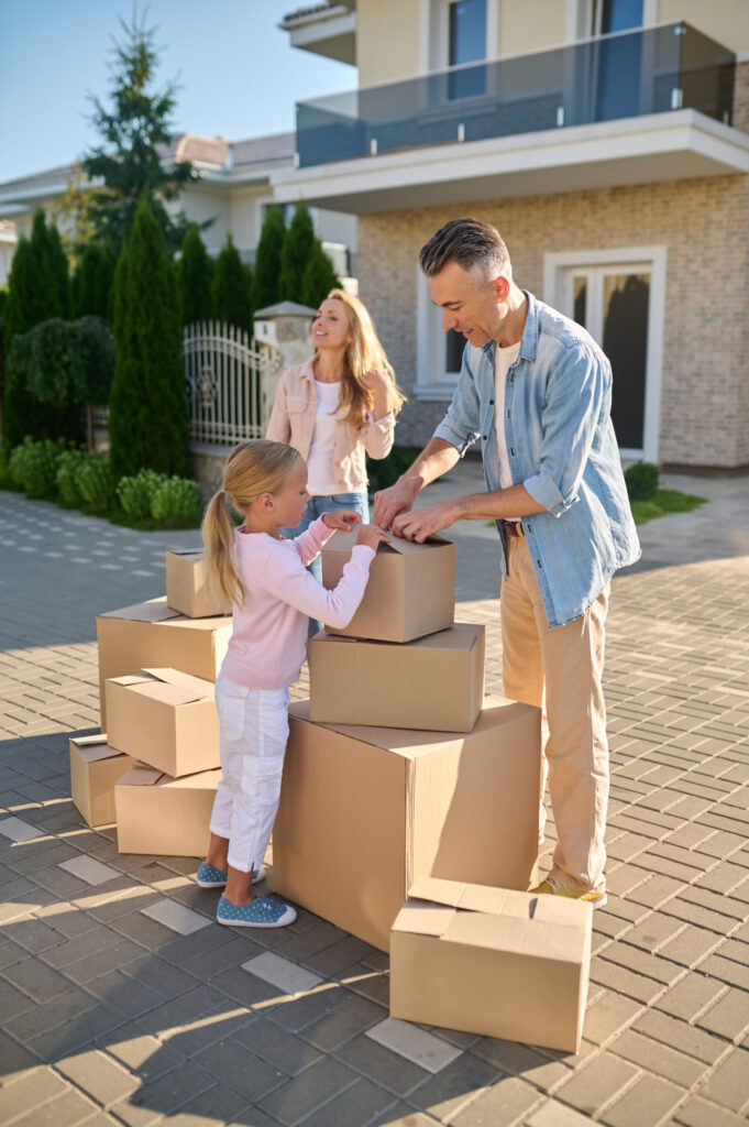 Famille préparant un déménagement longue distance: parent dépose un colis, enfants et autre parent souriants devant la maison avec des cartons.